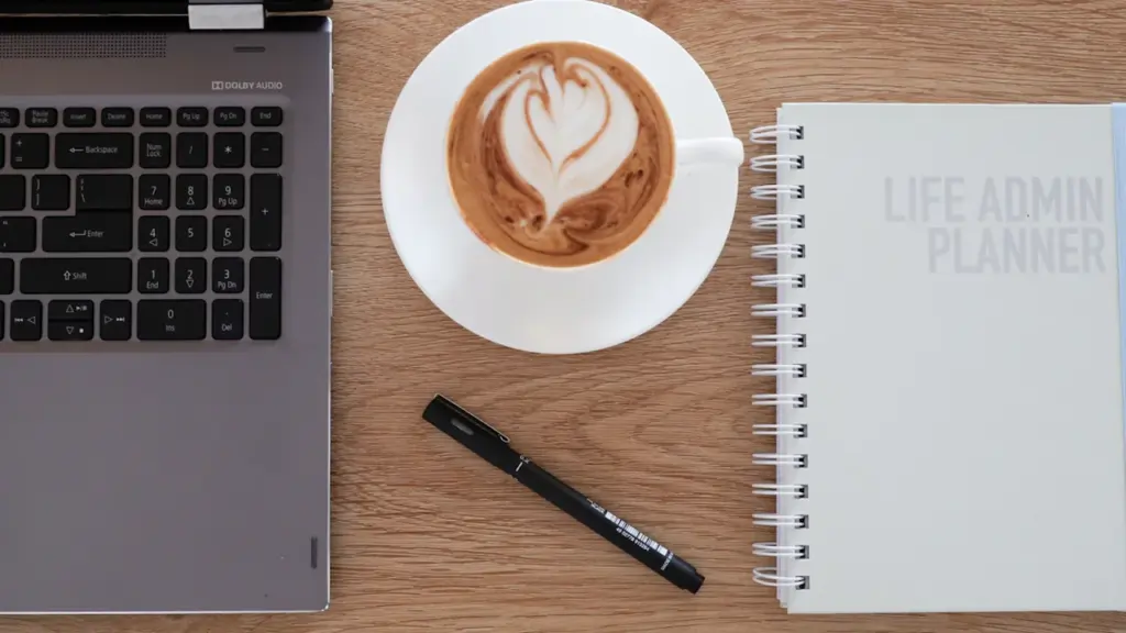 Ordenador portátil, taza de café y libreta sobre una mesa de madera clara.