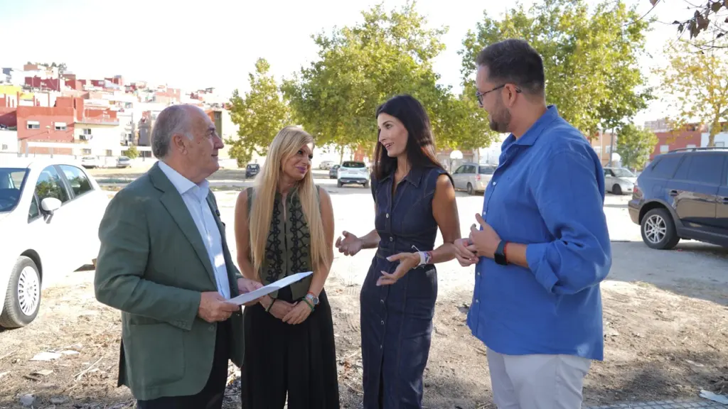 Cuatro personas conversan de pie en un descampado urbano con coches y edificios de fondo.